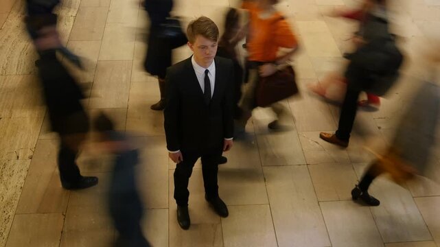 A man in a suit stands still in the midst of a moving crowd in a bustling urban location