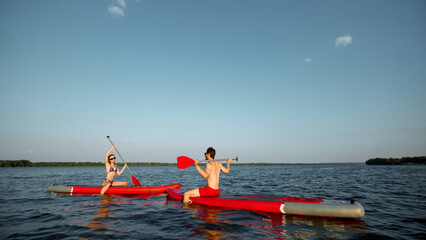 Naklejka premium Happy and relaxing young people, man and woman enjoying sunny evening on river, sailing on sup boards with paddles. Concept of sport, summer, active lifestyle, hobby, leisure time