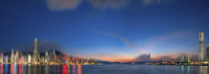 Naklejka premium A panoramic view of Hong Kong, China at dusk with a blurred cityscape and a calm harbor in the foreground.