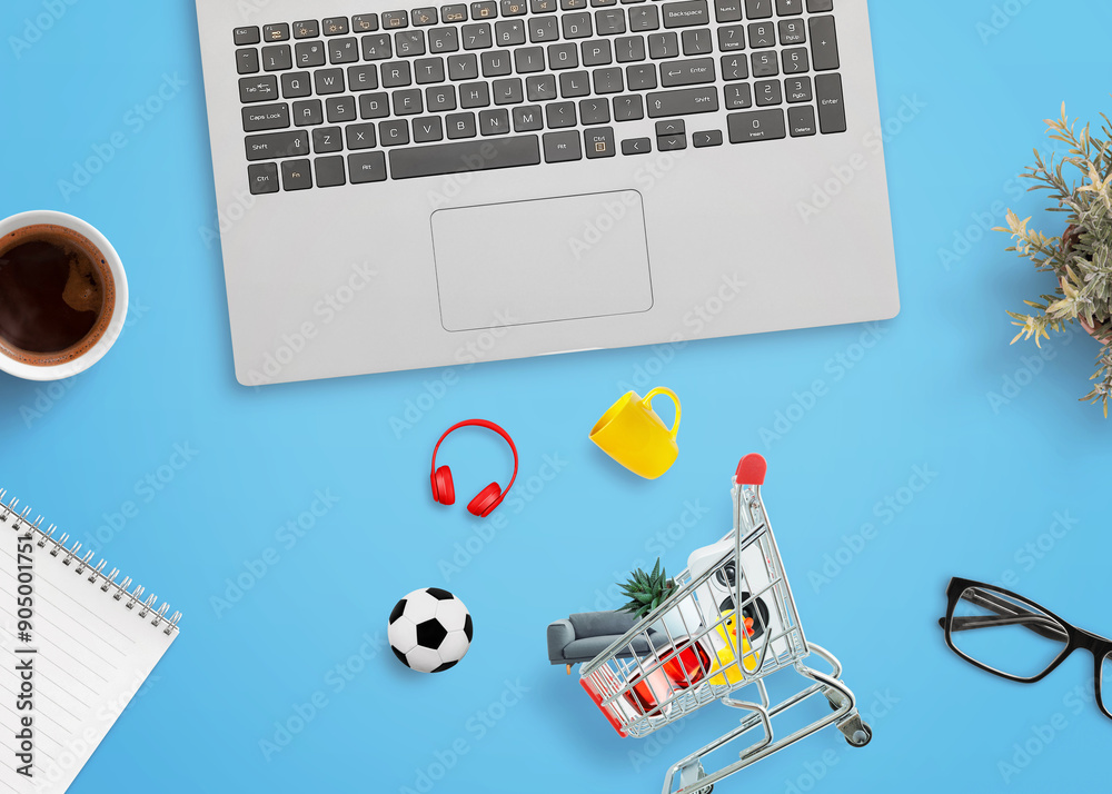 Poster Shopping cart on a desk with products around it and a laptop computer beside. Concept of online shopping and digital retail