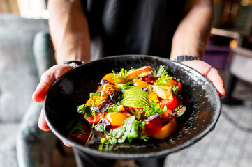 A person holding a black plate with a vibrant salad featuring greens, tomatoes, and avocado. The colorful and fresh ingredients make it perfect for culinary and healthy eating themes. 