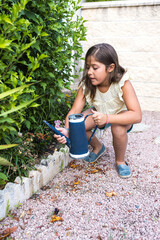 Black-haired girl watering plants in the garden with a blue watering can, squatting. Childhood, nature, outdoor, healthy, lifestyle, responsibility, home and cultivation.