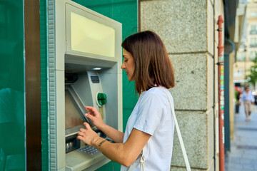 Young woman using an ATM on a city street, focused on the screen, with a green handbag.