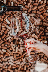 A hand with painted nails holds a glass as red wine is poured into it, with a background of wine corks. Perfect for themes of wine tasting, elegance, and celebration.