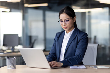 Businesswoman in formal attire working on laptop in modern office. Focused and diligent, she utilizes technology to accomplish tasks efficiently. Office environment reflects modern corporate setting.