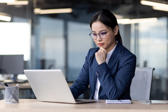Businesswoman concentrating on computer work while sitting at desk in modern office. Wearing glasses. Office environment, open workspace. Ideal for business, corporate, and professional concepts.