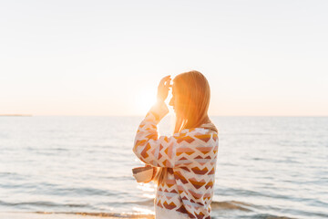 Silhouette side portrait of beautiful girl in cozy sweater holding cup of coffee. Young attractive woman enjoying a view of winter seaside shore with sun trail on the surface of romantic autumn ocean.