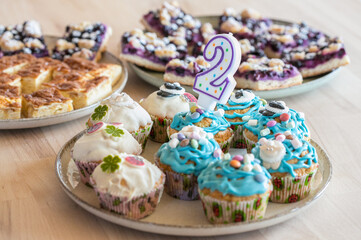 Cupcakes and fruit cake on table for 2nd birthday, blue and white frosting, apple cake, plum cake, candle in shape of number 2