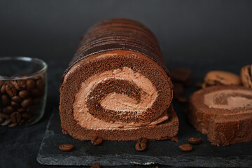 Chocolate roll with chocolate cream next to chocolate macaroons on a black tray next to coffee beans on a dark background