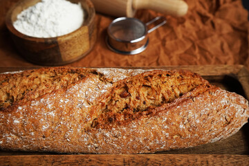 Fresh bread on a wooden tray and a brown napkin next to a bowl of flour and a wooden rolling pin on a brown background
