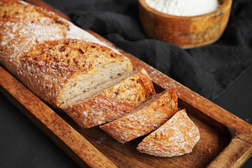 Fresh sliced ​​bread on a wooden tray next to a bowl of flour and a wooden rolling pin on a dark background