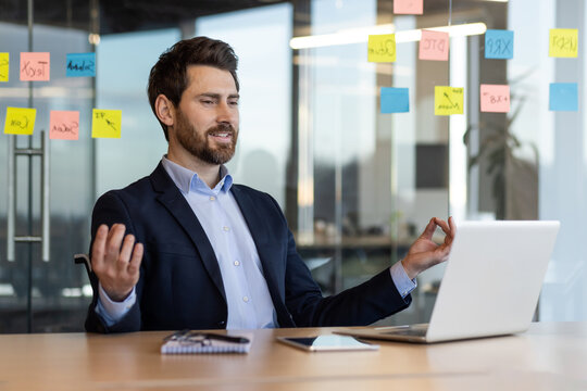 Businessman practicing mindfulness at office desk using laptop. Office worker managing stress through meditation. Concept of work-life balance in modern workplace with sticky notes on glass wall.