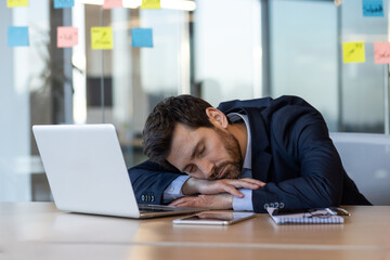 Exhausted businessman in suit sleeping on desk beside laptop and documents in modern office. Concept of workplace fatigue, overwork, stress, and work-life balance among office workers.