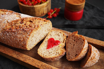 Fresh sliced ​​bread with a heart of berry jam on a wooden tray next to a bowl of red currants and jam on a dark background