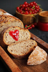 Fresh sliced ​​bread with a heart of berry jam on a wooden tray next to a bowl of red currants and jam on a dark background