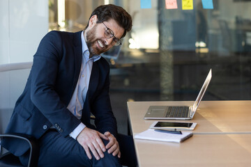 Businessman in formal attire experiencing leg pain while seated at desk in office environment. Man...