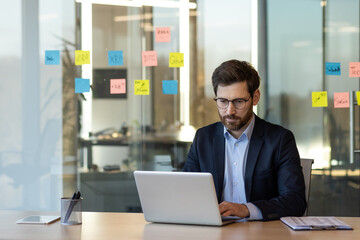 Focused businessman working on laptop in modern office. Glass wall with colorful sticky notes in background, reflecting collaborative and organized environment. Professional attire suggests corporate