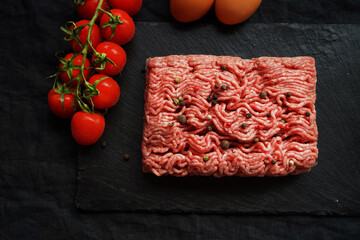 Minced meat with black and red peppers on a black slate tray next to a branch of tomatoes on a dark...