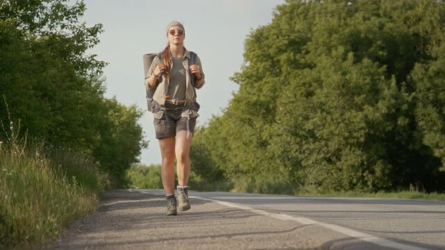 Full shot of young Caucasian woman with backpack and rolled-up sleeping mat walking along highway while traveling on vacation, looking back and sticking out thumb to hitch ride