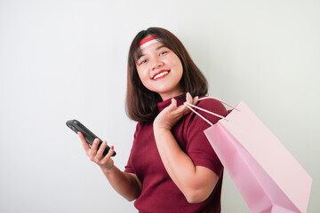 Naklejka premium Smiling young asian woman wearing Indonesian flag headband is looking her smartphone screen and carrying shopping bag, isolated over white background. Indonesian Independence Day.
