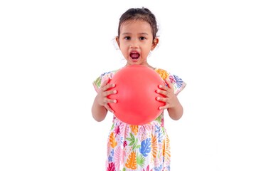 A cheerful little Asian girl in a colorful dress holding a red ball or balloon with both hands on a...