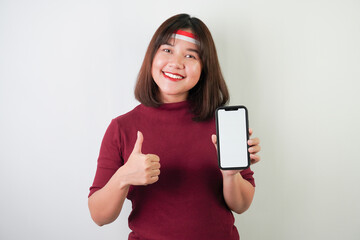 Fototapeta premium Smiling young asian woman wearing Indonesian Flag headband is showing vertical blank smartphone screen for advertising with okay gesture, isolated over white background. Indonesian Independence Day.