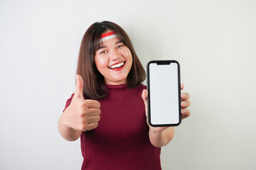 Fototapeta premium Smiling young asian woman wearing Indonesian Flag headband is showing vertical blank smartphone screen for advertising with okay gesture, isolated over white background. Indonesian Independence Day.