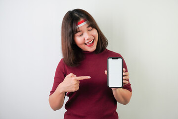 Fototapeta premium Smiling young asian woman wearing Indonesian Flag headband is showing vertical blank smartphone screen for advertising, isolated over white background. Indonesian Independence Day.