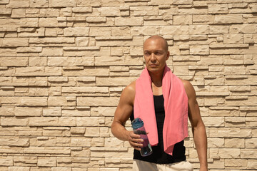 Obraz premium Portrait of athlete adult man with pink towel and water bottle tired after training against beige wall. Summer outdoors workout. Copy space.