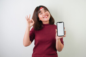 Fototapeta premium Smiling young asian woman wearing Indonesian Flag headband is showing vertical blank smartphone screen for advertising with okay gesture, isolated over white background. Indonesian Independence Day.