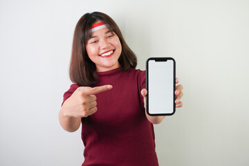 Fototapeta premium Smiling young asian woman wearing Indonesian Flag headband is pointing vertical blank smartphone screen for advertising, isolated over white background. Indonesian Independence Day.
