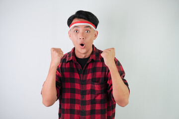 Excited young asian man wearing Indonesian flag headband, smiling expression, hands raised with clenched fist gesture, isolated over white background. Concept for Indonesian Independence Day.