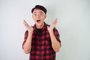 Excited young asian man wearing Indonesian flag headband, smiling expression, hands raised with open hand palm gesture, isolated over white background. Concept for Indonesian Independence Day.