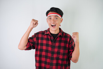 Excited young asian man wearing Indonesian flag headband, smiling expression, hands raised with clenched fist gesture, isolated over white background. Concept for Indonesian Independence Day.