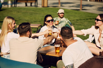 Joyful gathering. Group of friends, men and women sitting outdoors in restaurant, talking, laughing and drinking refreshing beer. Concept of summer, leisure, friendship, meeting, fun, relaxation