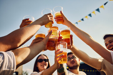 Group of people, friends gathering on picnic, raising glasses with beer, clinking under blue sunny...