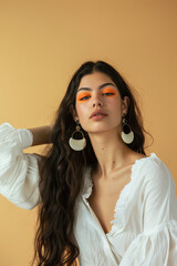 A young woman with long dark hair and vibrant orange eye makeup, wearing a white blouse and large earrings, posing with a serene expression against a beige background