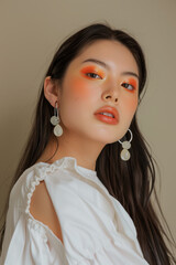 A young woman with long dark hair and vibrant orange eye makeup, wearing a white blouse and large earrings, posing with a serene expression against a beige background