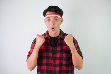 Excited young asian man wearing Indonesian flag headband, smiling expression, hands raised with clenched fist gesture, isolated over white background. Concept for Indonesian Independence Day.