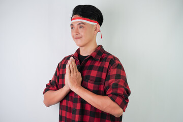 Fototapeta premium Excited young asian man wearing Indonesian flag headband, smiling expression with hands pressed together gesture, isolated over white background. Concept for Indonesian Independence Day.