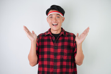 Surprised young asian man wearing Indonesian flag headband, amazed expression with hands open gesture, isolated over white background. Concept for Indonesian Independence Day.