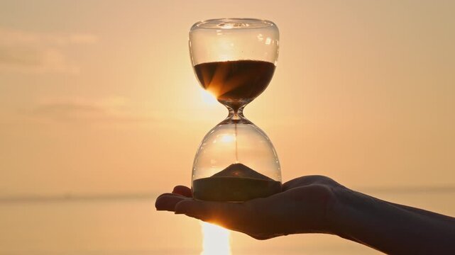 Hourglass concept time woman holding glass hourglass in palm on blurred background of golden sun disk sky in sunset rays in summer close-up. Golden sand pours quickly. Time concept sand. Relax