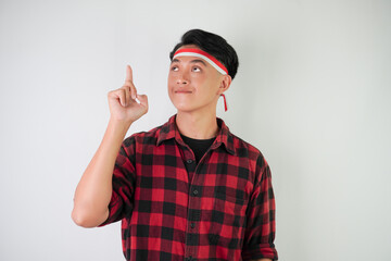 Smiling young asian man wearing Indonesian flag headband and pointing fingers on copy space upside, isolated over white background. Concept for Indonesian Independence Day.