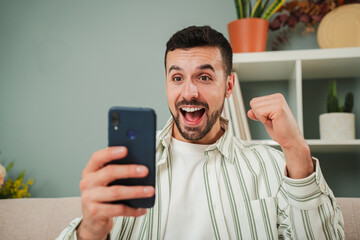 Young man celebrating that he has won a sports bet while checking the result on a smartphone app. A happy guy betting at an online casino with a cellphone. An excited male watching the lottery prize
