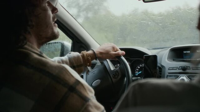 Medium close-up rear shot of young Caucasian man and woman driving in car together in pouring rain, on way to vacation, having lively conversation, looking at each other and smiling