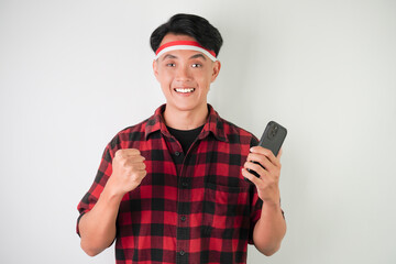 Surprised shocked young asian man wearing Indonesian Flag headband is watching vertical smartphone screen for advertising, isolated over white background. Indonesian Independence Day.