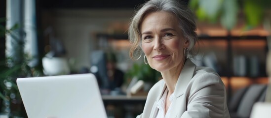 Fototapeta premium Successful senior businesswoman working on laptop, smiling confidently in modern office. Concept of older women in business, career woman, leadership, technology, and entrepreneurship