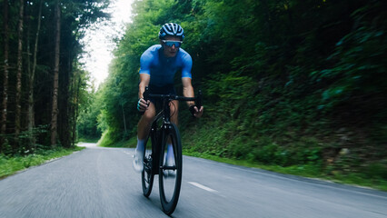 Male road racing cyclist in a blue sports jersey, with helmet and glasses, sprinting up a hill...