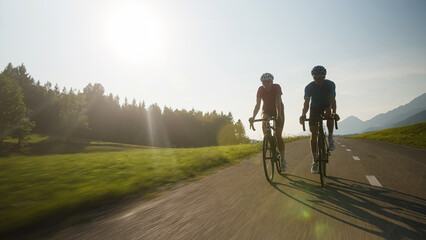 Obraz premium Front view of two people, a male, and female racing cyclists ride on a paved road through beautiful mountain nature at sunset
