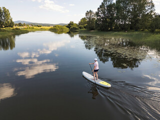 Woman paddling sup board, enjoying tranquil moments on mountain lake surrounded by serene nature, aerial shot. Balanced lifestyle and wellbeing concepts.